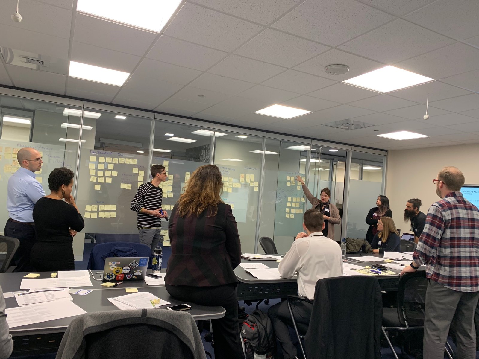 A workshop in progress. Participants stand at a glass wall covered in sticky notes grouped under headings including Measurement and Metrics, Data Ecosystem and Tools, Hiring, and Reporting, while others work at tables covered in printed materials.